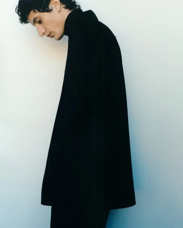 Profile portrait of an adult male model wearing a black turtleneck sweater against a white backdrop, minimalist fashion and studio lighting.