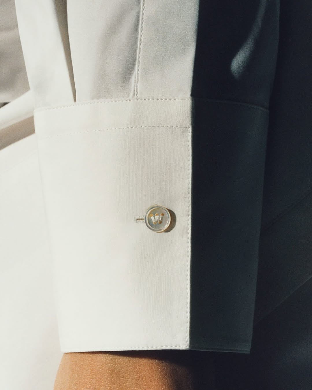 Close-up on a white dress shirt cuff with a round cufflink featuring the letter 'W,' partially shadowed, highlighting the fabric texture.