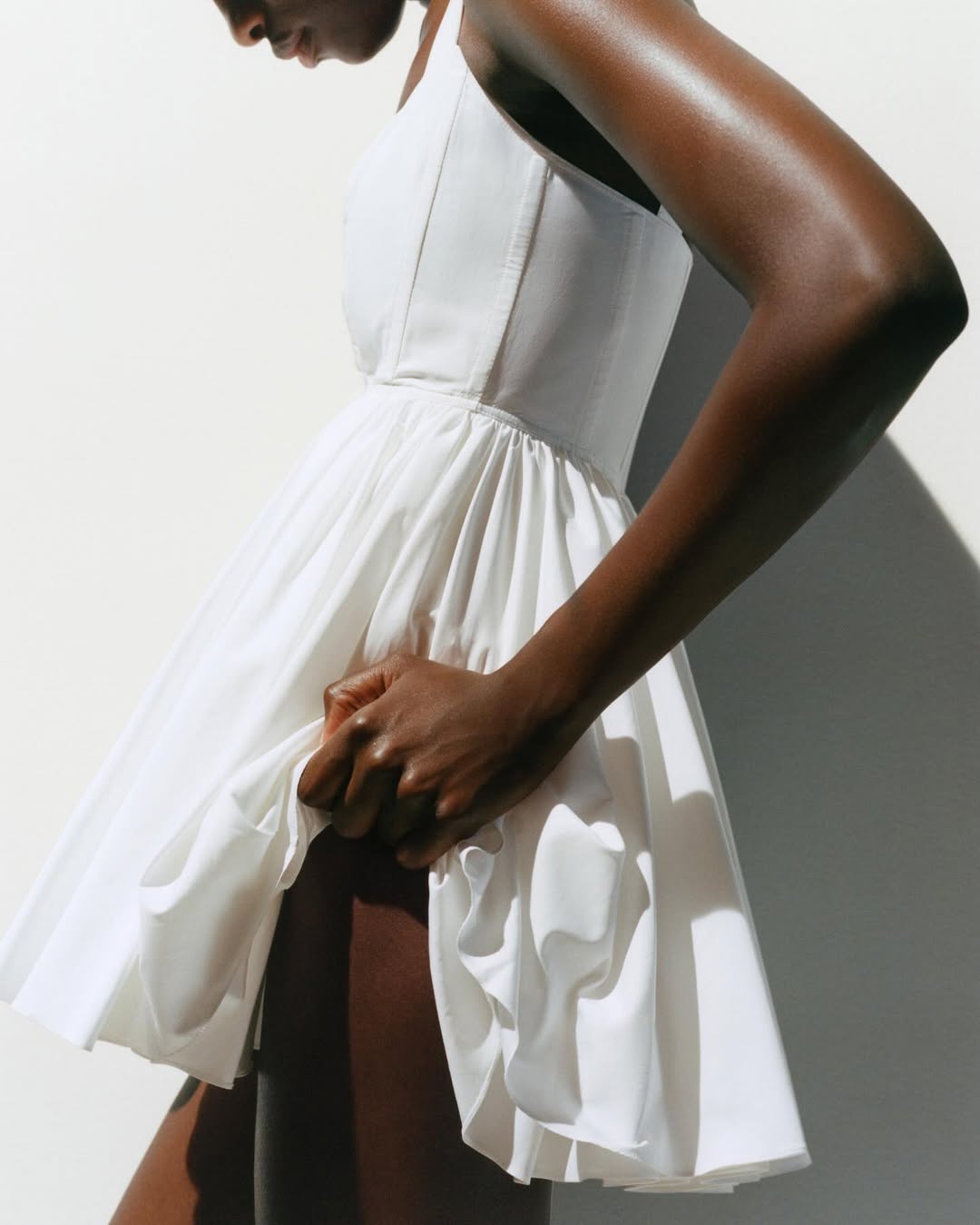 An adult female model with dark skin poses indoors wearing a short white dress in a fashion editorial shot against a white wall.