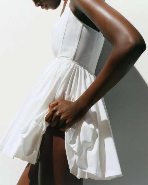 An adult female model with dark skin poses indoors wearing a short white dress in a fashion editorial shot against a white wall.