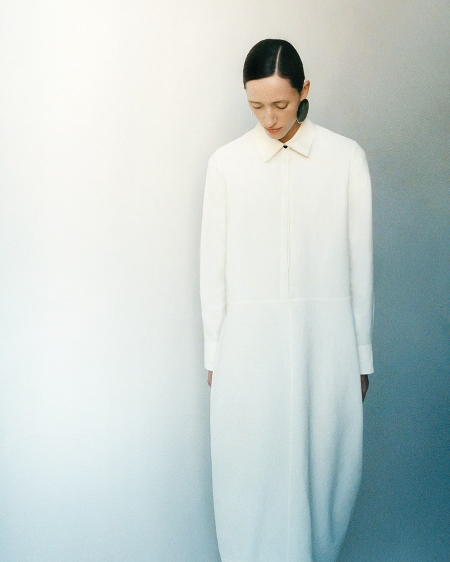 Model with slicked back hair wearing a long white button-down dress standing against a gradient wall.