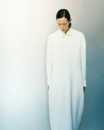 Model with slicked back hair wearing a long white button-down dress standing against a gradient wall.