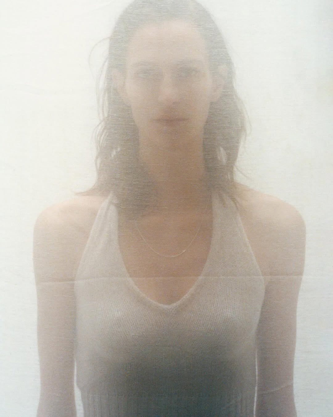 Portrait of a young adult woman with light skin and long hair, wearing a halter top and necklace, set against a soft, hazy white background.