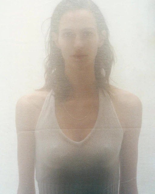 Portrait of a young adult woman with light skin and long hair, wearing a halter top and necklace, set against a soft, hazy white background.