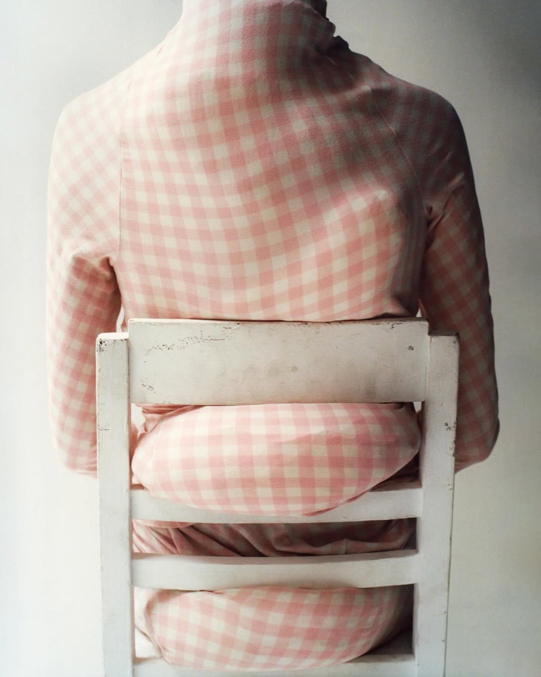 Back view of a person in a pink and white checkered top, seated on a weathered white wooden chair, against a plain white background.