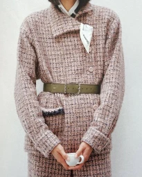 Model wearing a pink tweed jacket and skirt belted with a green olive belt, holding a small white ceramic cup.
