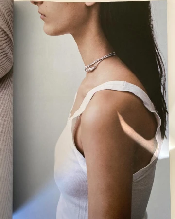 Close-up profile of a woman wearing a white tank top and a delicate silver choker necklace against a pale neutral background.
