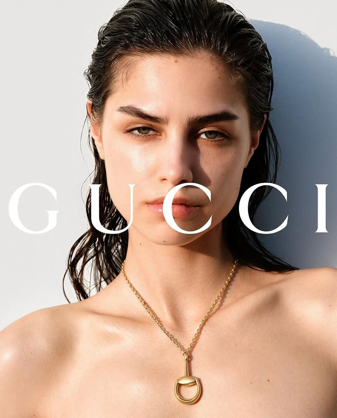 Close-up fashion portrait of a young adult woman with dark hair, wearing a gold necklace, bright studio lighting, neutral white background.