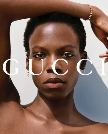 Close-up portrait of a Black woman with short dark hair wearing gold horsebit earrings and a matching bracelet against a light grey background.