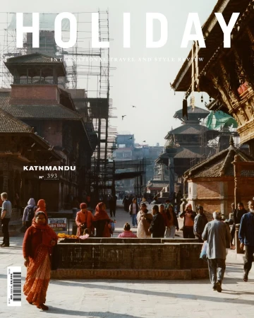 Magazine cover featuring a bustling Kathmandu street scene with people, historic architecture, and scaffolding against a bright sky.