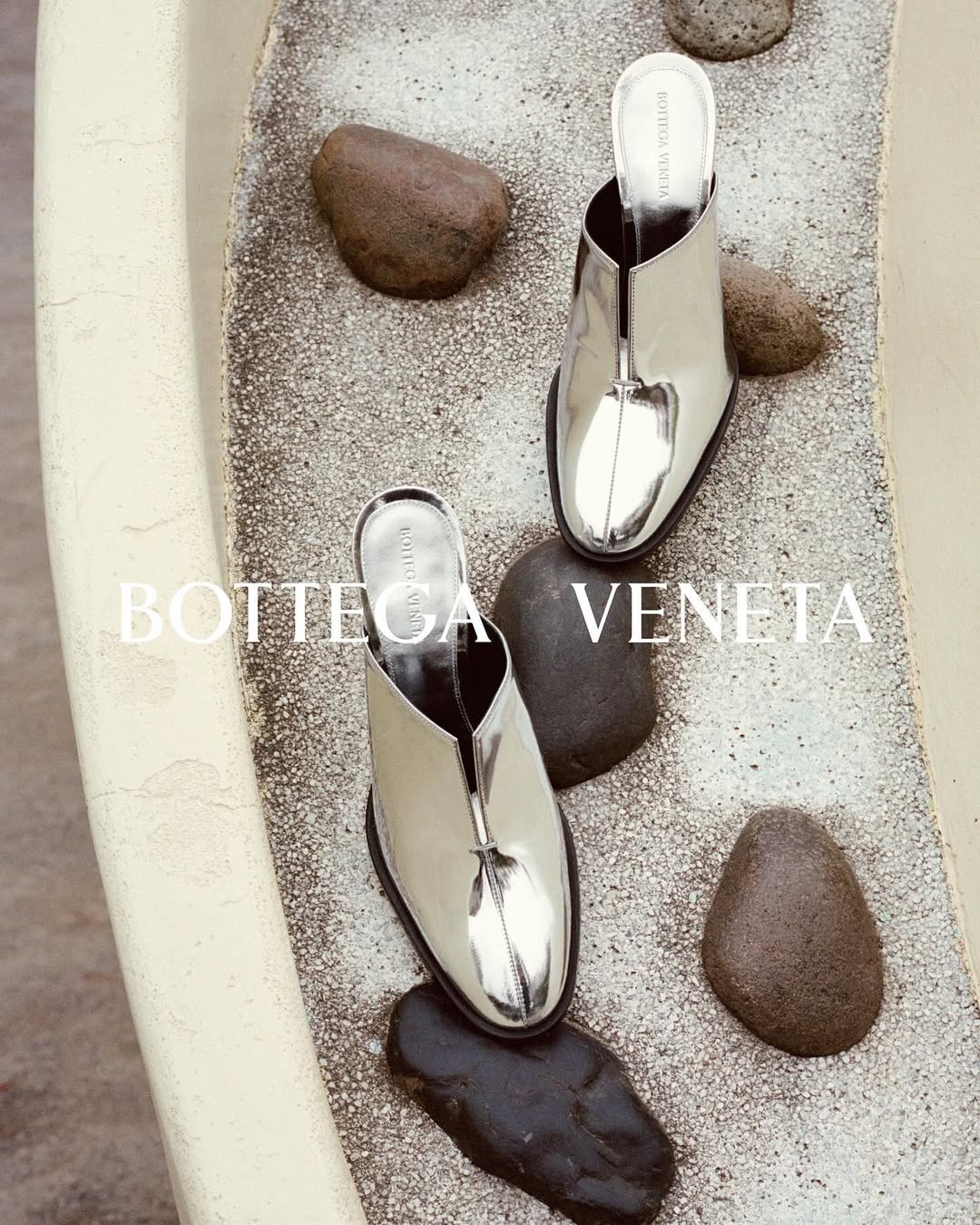 Two silver Bottega Veneta mule heels are displayed on a speckled surface with smooth dark stones, creating a fashion editorial shot.