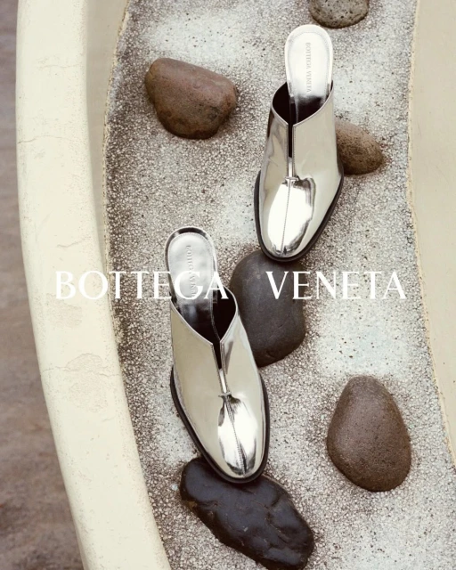 Two silver Bottega Veneta mule heels are displayed on a speckled surface with smooth dark stones, creating a fashion editorial shot.