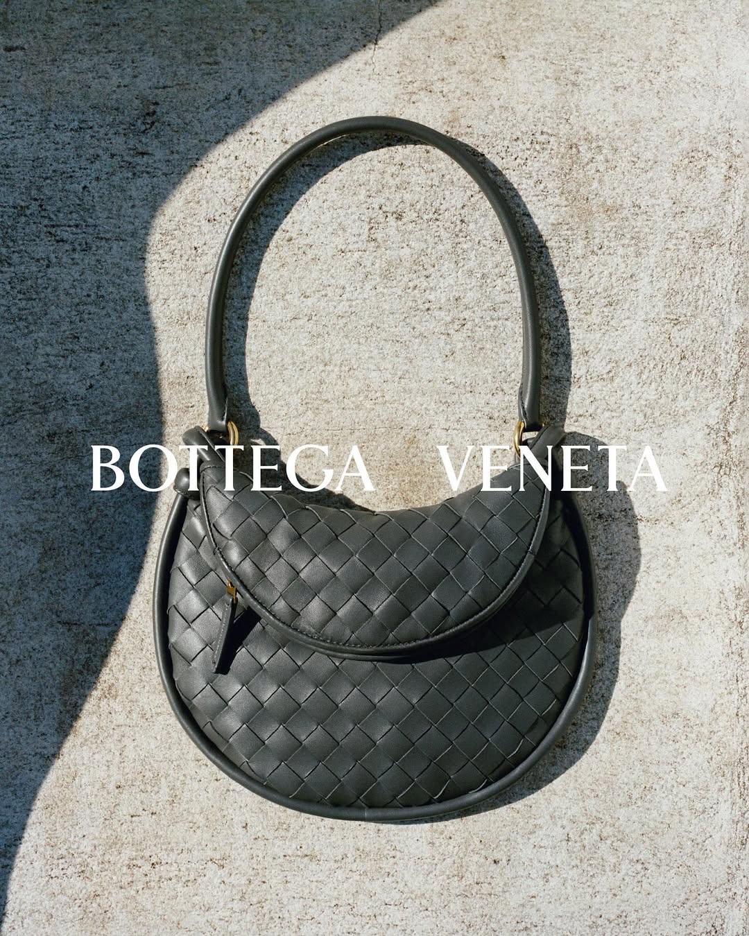 Gray Bottega Veneta woven handbag with top handle, displayed on a textured gray surface with dappled sunlight, fashion accessory.