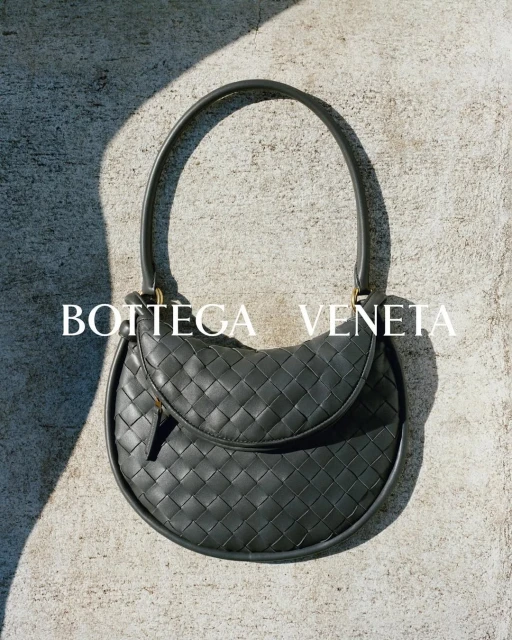 Gray Bottega Veneta woven handbag with top handle, displayed on a textured gray surface with dappled sunlight, fashion accessory.