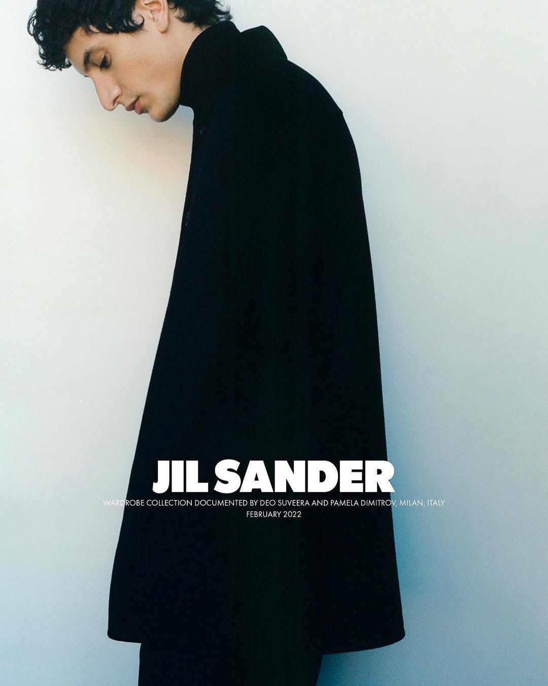 Fashion shot of a young adult male model wearing a black coat with a high collar, posing indoors against a plain light-colored wall.