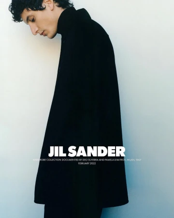 Fashion shot of a young adult male model wearing a black coat with a high collar, posing indoors against a plain light-colored wall.