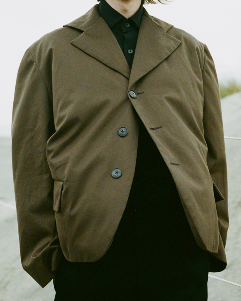 Person in a brown blazer and black shirt, standing outside in a neutral background with soft lighting.