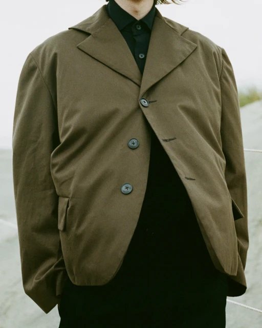 Person in a brown blazer and black shirt, standing outside in a neutral background with soft lighting.