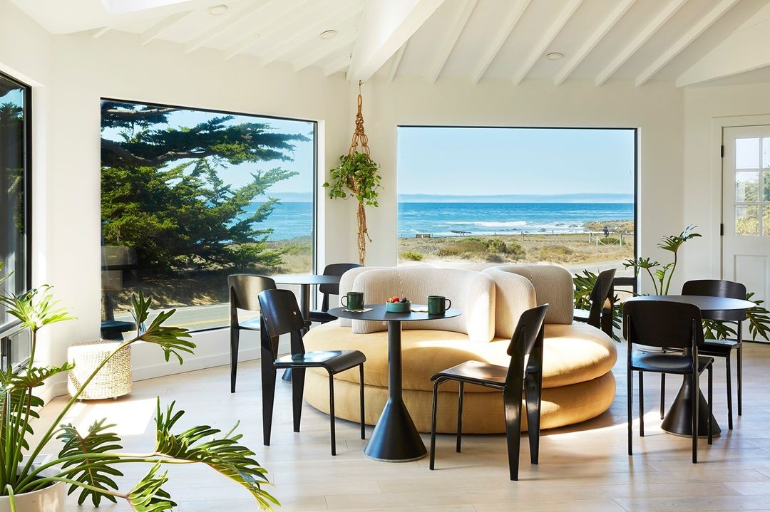 Bright, modern indoor lounge area with large view windows, round sofa, black chairs, and tables with ocean view background.