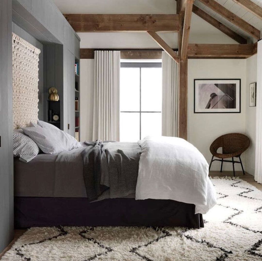 Bedroom interior with a queen-size bed featuring a woven headboard, neutral bedding, patterned rug, wooden beams, and soft, natural light.