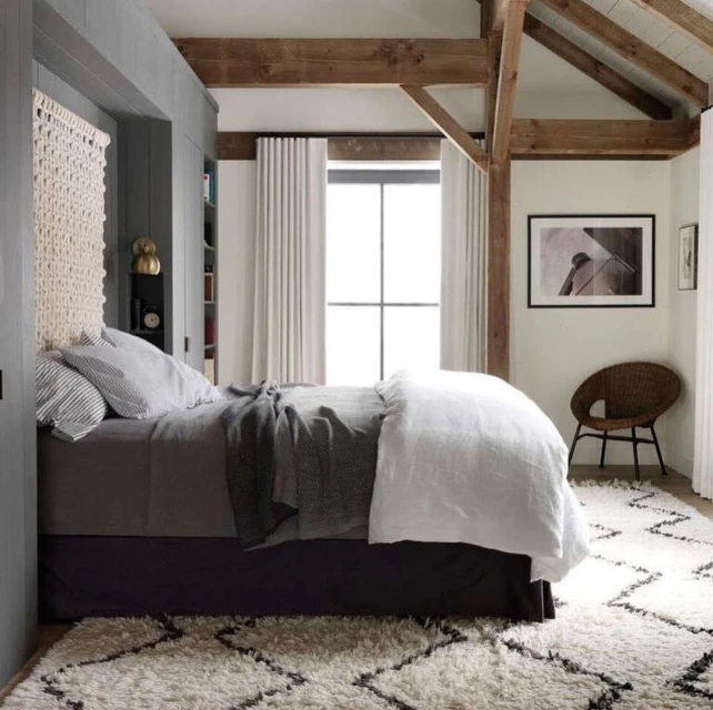 Bedroom interior with a queen-size bed featuring a woven headboard, neutral bedding, patterned rug, wooden beams, and soft, natural light.