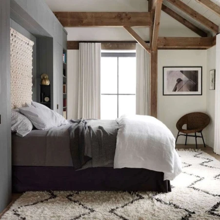 Bedroom interior with a queen-size bed featuring a woven headboard, neutral bedding, patterned rug, wooden beams, and soft, natural light.