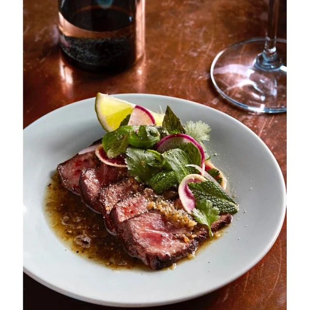 Gourmet plate with sliced steak, a lime wedge, and herbs including cilantro and mint served on a light blue plate with dark sauce.
