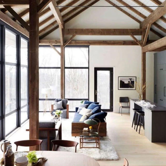 Living room interior with wooden beams, large windows overlooking nature, sofas, chairs, tables, and a kitchen island with stools.