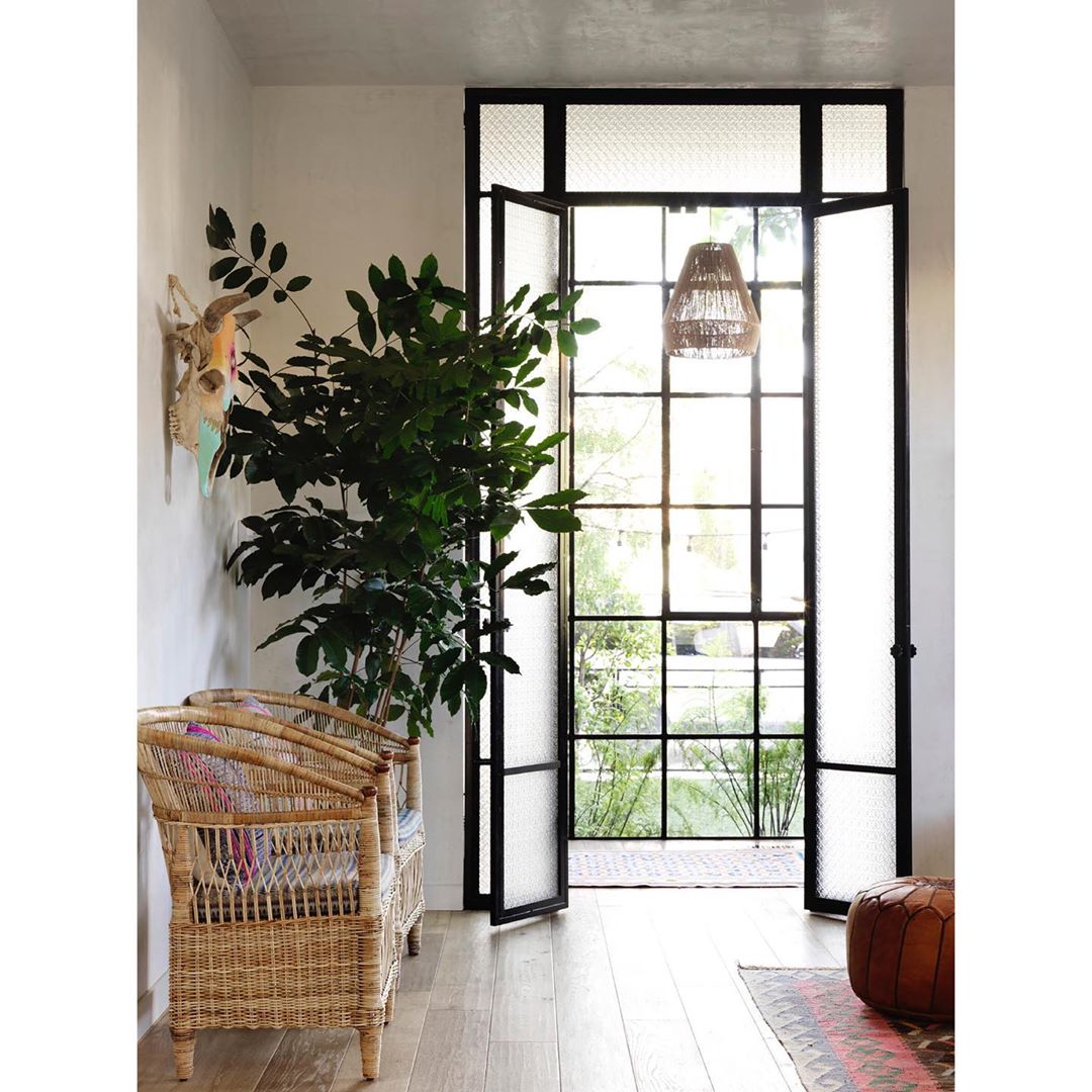Living room interior with wicker chairs, potted plant, black-framed glass doors to garden, woven pendant light, wood floor, and pouf.