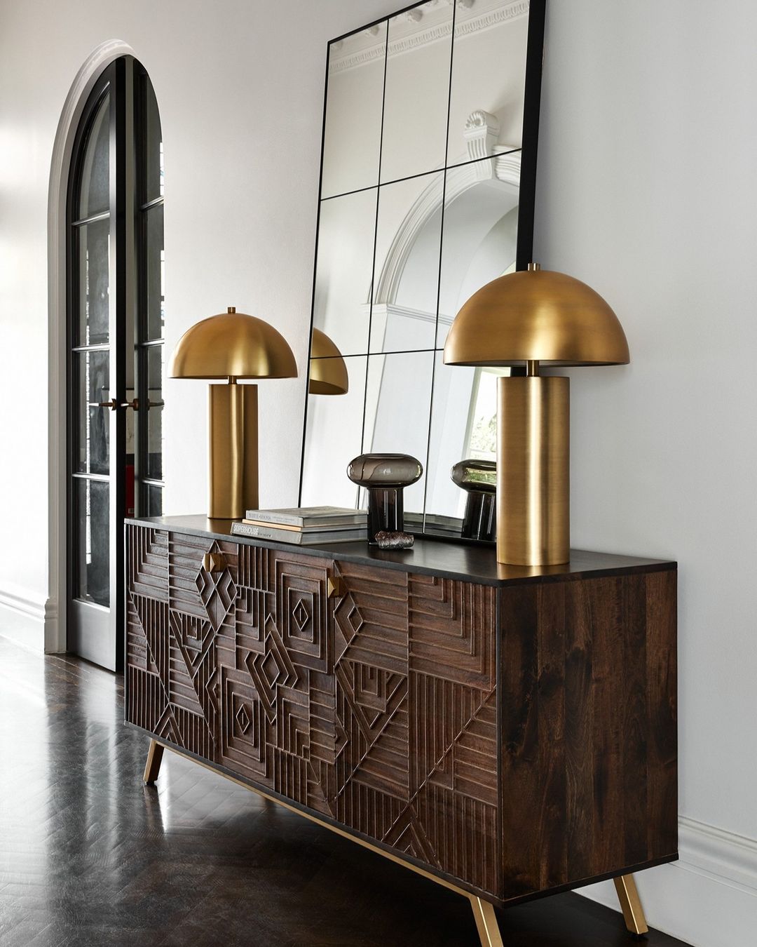 Interior design featuring a dark wood console table with gold accents, topped with gold dome lamps, against a mirrored wall.