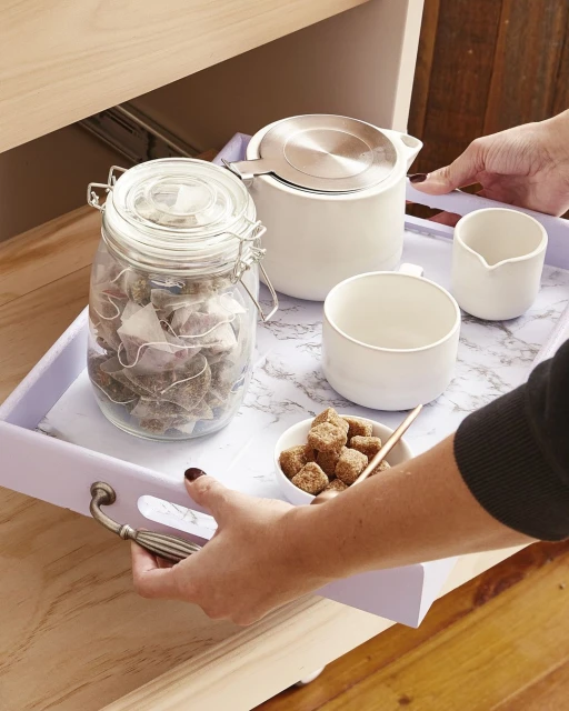 Overhead shot of a light-purple tray holding tea in a white ceramic teapot, sugar bowl, milk jug, teacup, and tea bags in a glass jar.
