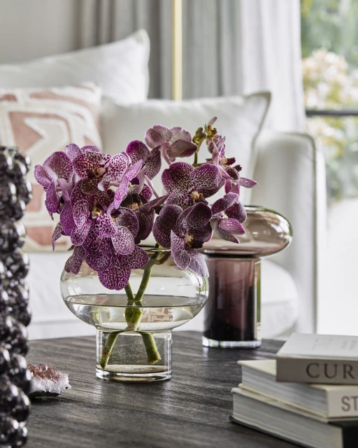 Floral arrangement in a clear vase with purple orchids on a dark wood table, soft white couch with pillows in the background.