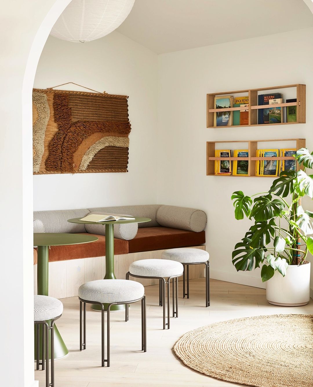 Stylish seating area with olive green tables, cushioned bench, round stools, shelving with books, houseplants, and a jute rug.