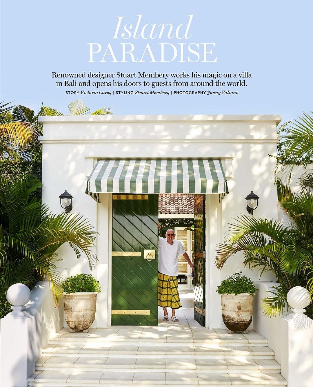 Mature man in a yellow sarong stands smiling in the doorway of a white villa surrounded by tropical plants on a sunny day.