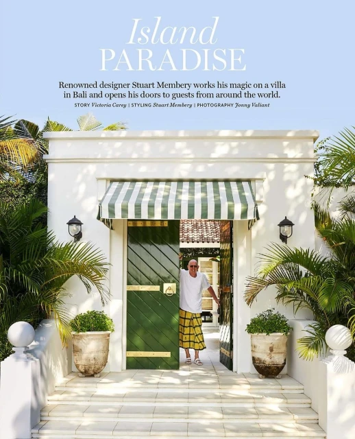 Mature man in a yellow sarong stands smiling in the doorway of a white villa surrounded by tropical plants on a sunny day.
