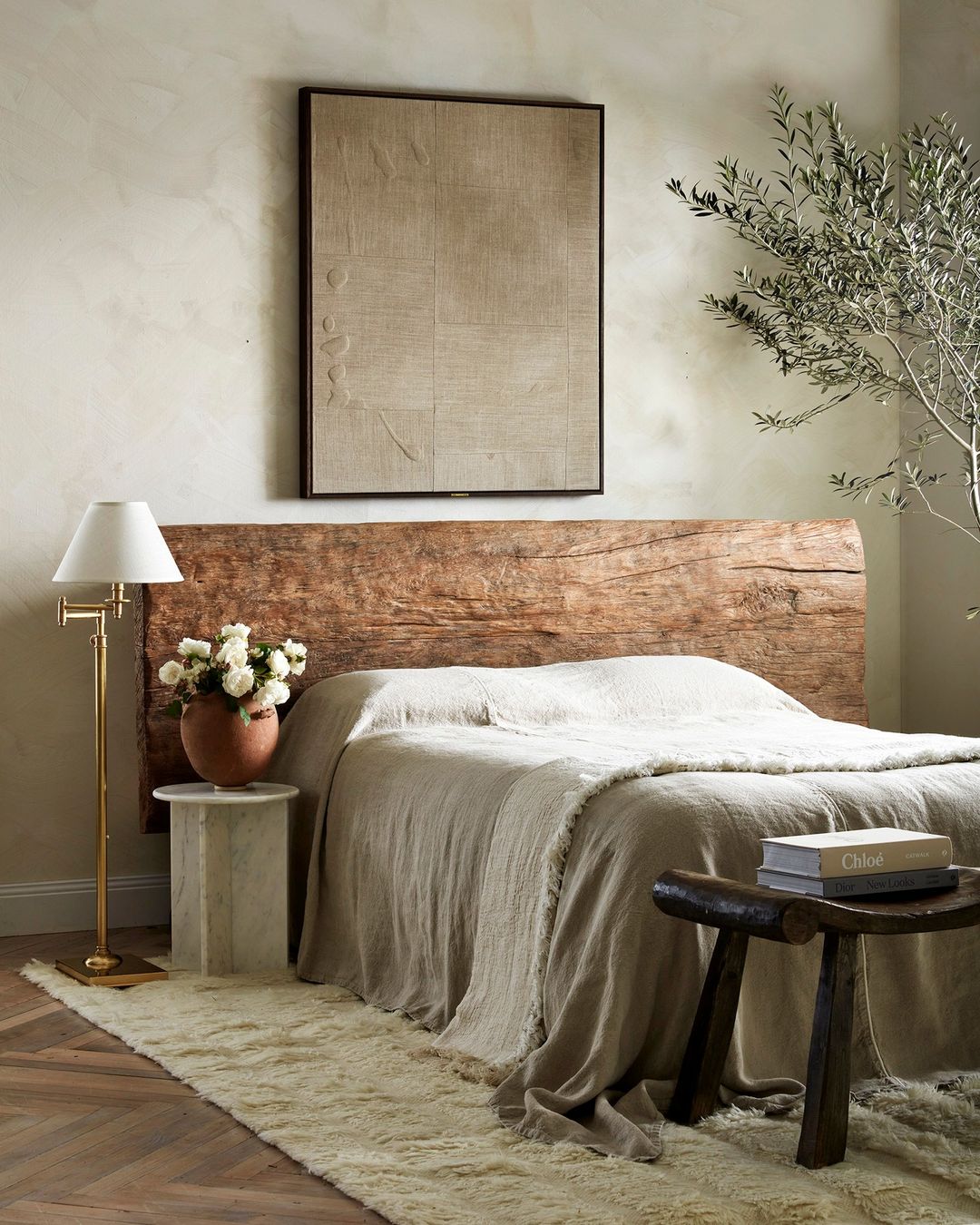 Bedroom interior with a wooden headboard, neutral bedding, side table with flowers, lamp, and olive tree branch creating a calming ambiance.