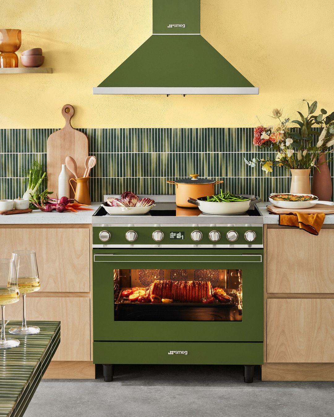 Olive green Smeg range cooker with overhead vent, pots of food, and countertop vegetables in a residential kitchen setting.
