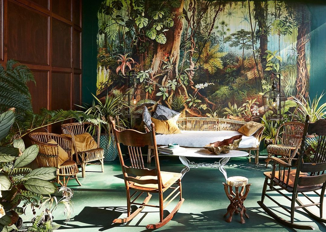 Living room with vintage furniture, including chairs, a sofa, table, and indoor plants in front of a forest-themed mural.
