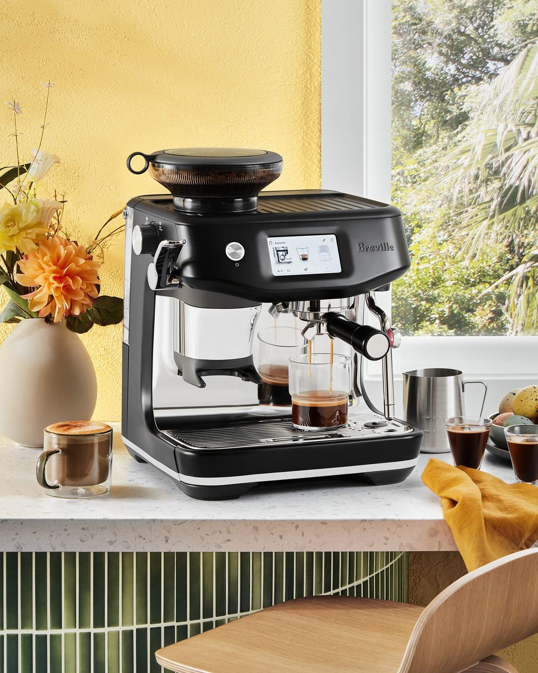 Modern black espresso machine brews coffee into glass cups on a marble countertop, with fresh flowers and light yellow wall in background.