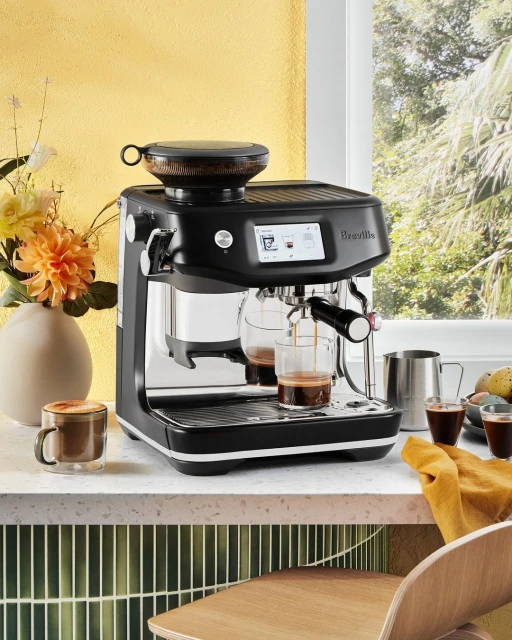 Modern black espresso machine brews coffee into glass cups on a marble countertop, with fresh flowers and light yellow wall in background.