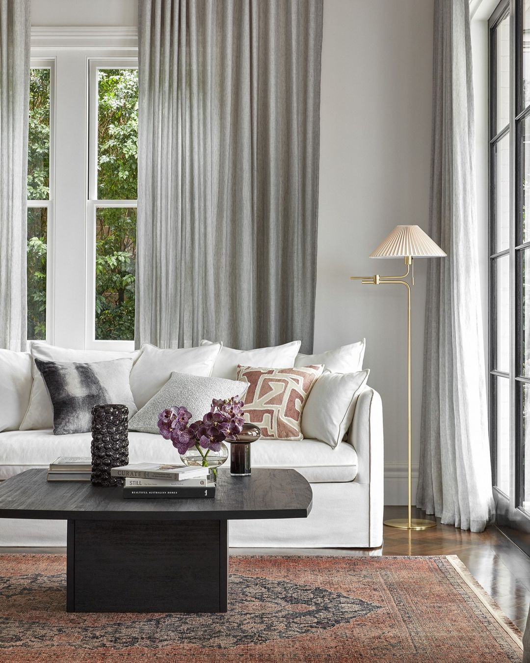Living room interior with a white couch, dark wood coffee table, flowers, books, a gold lamp, and a patterned rug in neutral tones.