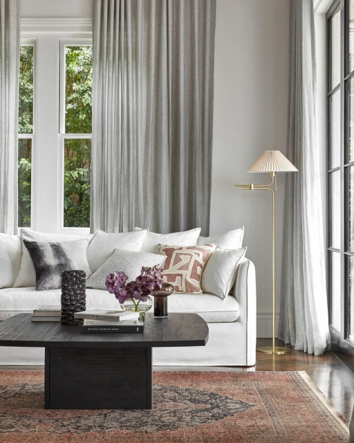Living room interior with a white couch, dark wood coffee table, flowers, books, a gold lamp, and a patterned rug in neutral tones.