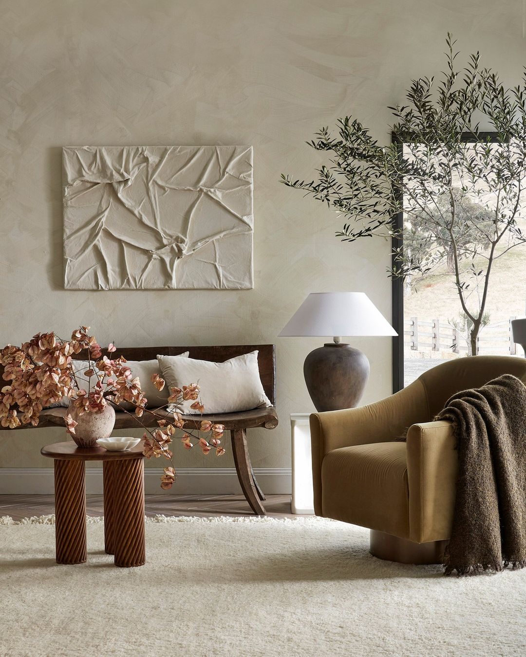Interior design featuring a tan armchair with throw blanket, a wooden bench with pillows, textured wall art, and an olive tree branch.