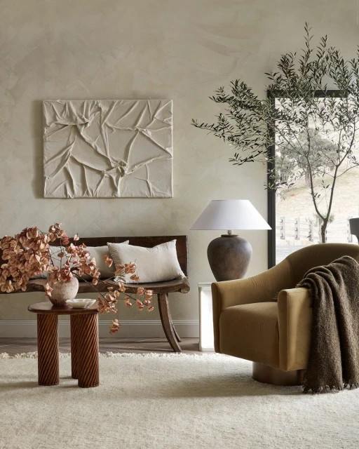 Interior design featuring a tan armchair with throw blanket, a wooden bench with pillows, textured wall art, and an olive tree branch.