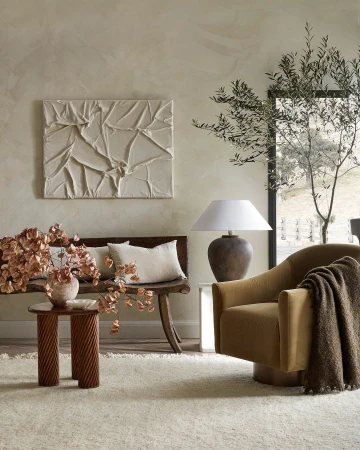 Interior design featuring a tan armchair with throw blanket, a wooden bench with pillows, textured wall art, and an olive tree branch.