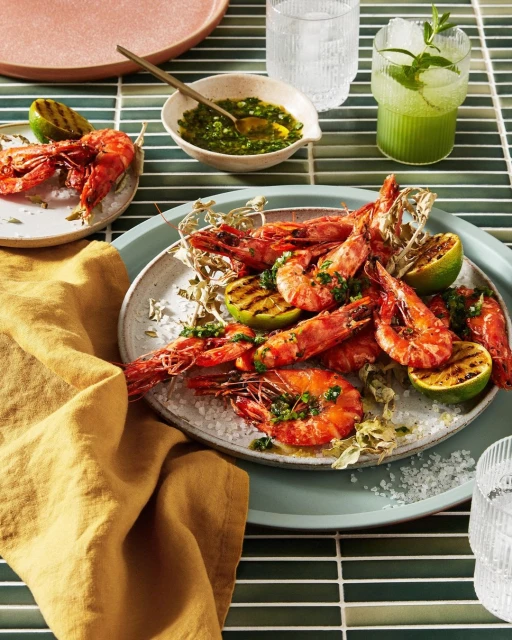 Overhead shot of grilled shrimp and lime wedges served on a light blue plate with dipping sauce and beverages on a tiled surface.