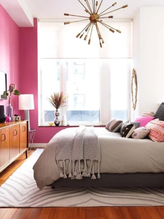 Bedroom interior with a bed, pillows, and a stylish chandelier, featuring pink wall, neutral bedding, and hardwood floor.