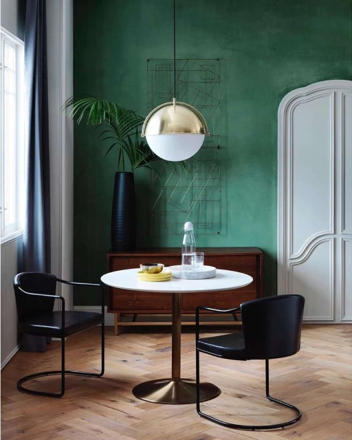 Interior of dining area with a round table and two black chairs, lit by a modern pendant light, set against a green wall and wooden floor.