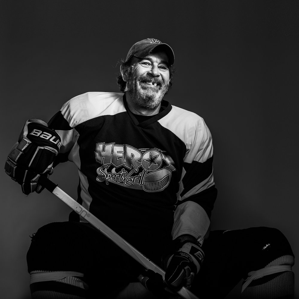 Monochrome portrait of a man in a hockey jersey and baseball cap, smiling with missing teeth, holding a hockey stick against a grey backdrop.