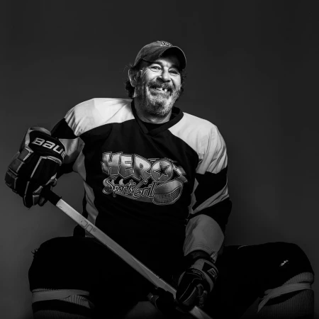 Monochrome portrait of a man in a hockey jersey and baseball cap, smiling with missing teeth, holding a hockey stick against a grey backdrop.